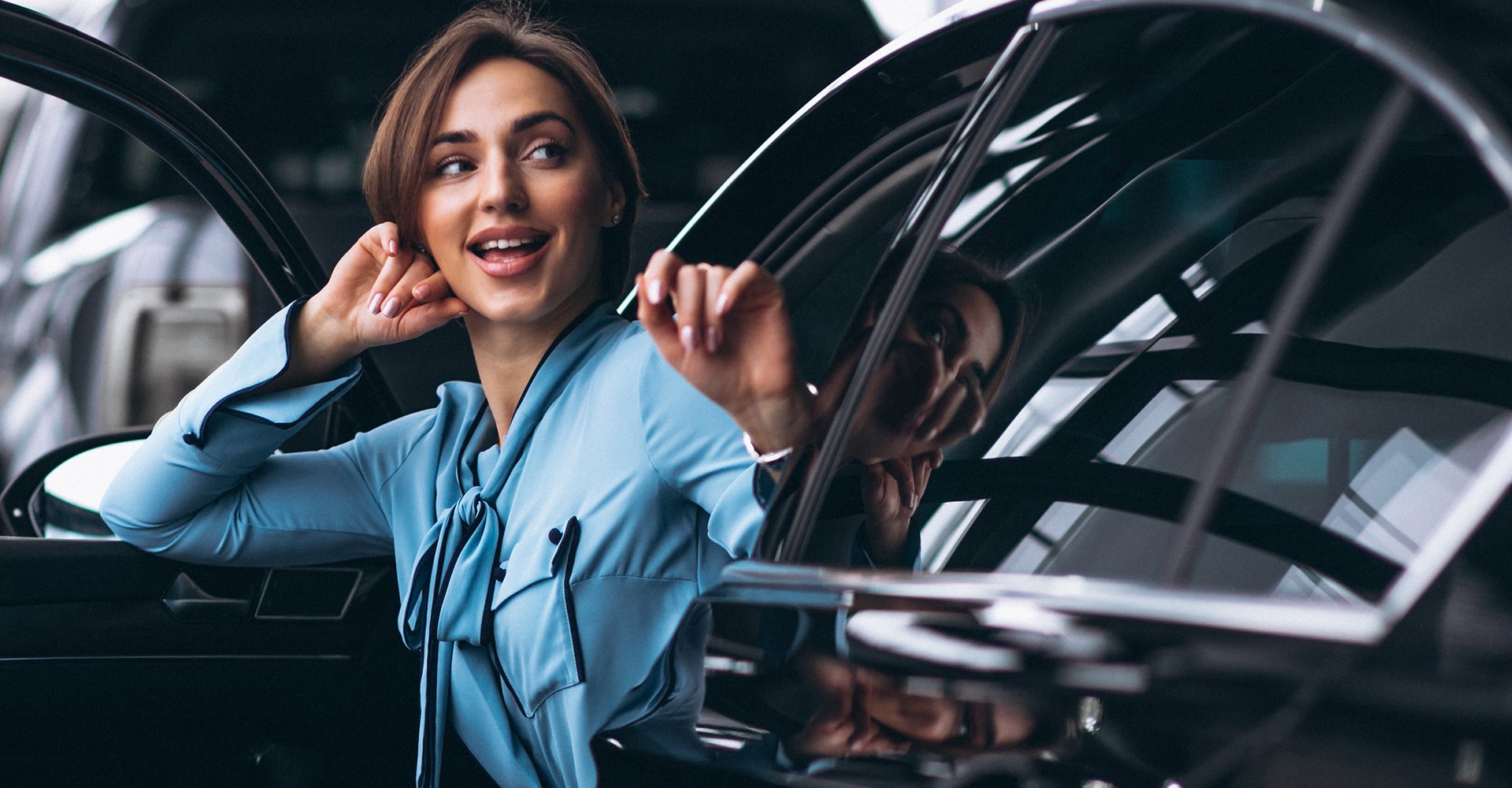 Woman in a light blue blouse smiling and gesturing from inside a shiny black car, reflecting the quality and attention to detail of Mr.Shine's car wash services.
